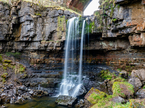 View of Ashgill Force waterfall cascading over layered rock formations with mossy stones and a stone bridge above in Alston, England, United Kingdom.