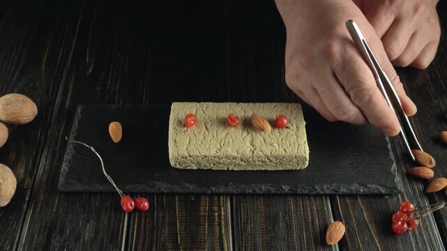 Hands carefully place almonds and red berries on a rectangular green dish using tweezers, with scattered almonds and berries on a dark wooden table, creating an artistic food presentation