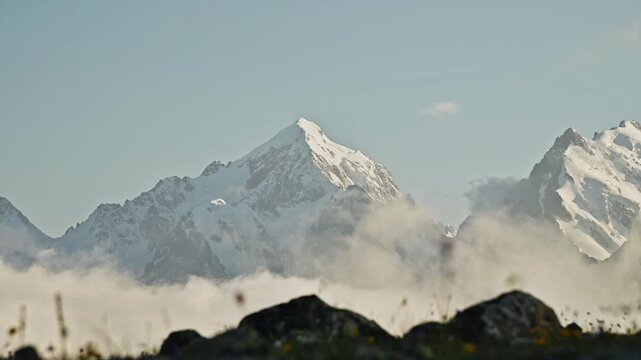 Snow-capped mountains emerge from clouds with rocky foreground in serene landscape