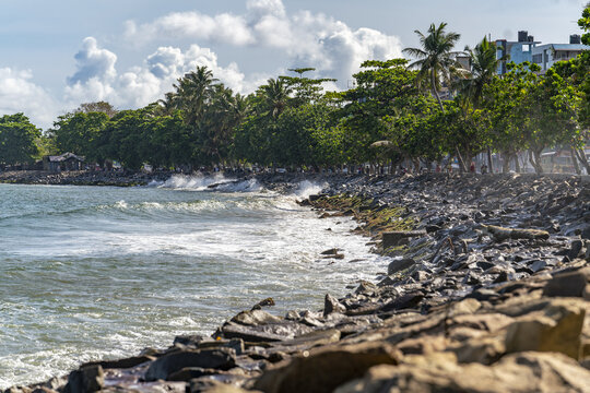 View of the Marine Walk with rocky breakwaters, crashing waves, and lush trees along the coastline in Galle, Southern Province, Sri Lanka.