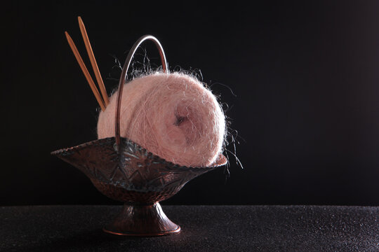 Pink ball of fluffy yarn and wooden knitting needles in metal vase on black background. Two wooden knitting needles are sticking out of vase. Image is ideal for advertising luxury yarns. Copy space