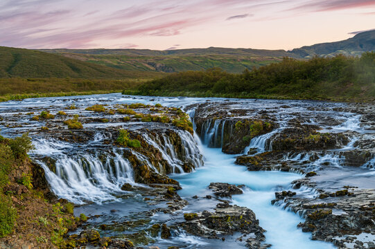 View of the cascading turquoise waters of a waterfall flowing over dark volcanic rock surrounded by lush green hills under a soft sunset sky in Iceland.