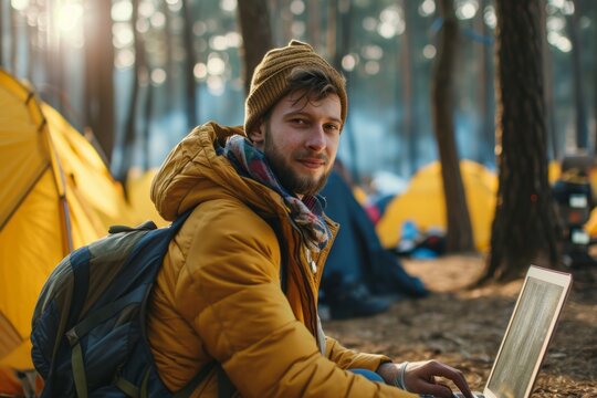 Young man with beard wearing yellow jacket and knitted hat, working on laptop outdoors in forest campsite, surrounded by colorful tents and nature, showcasing remote work lifestyle
