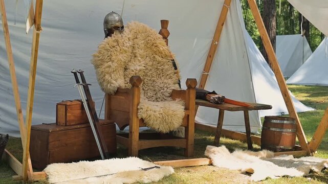 A medieval reenactor sits on a wooden chair next to various historical props, including weapons and a wooden chest. The setting features tents and natural surroundings, suggesting a historical camp