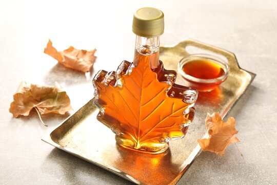 Leaf shaped bottle of maple syrup, bowl and dried leaves on light grey table, closeup