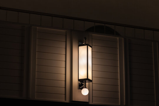 A warm interior wall lantern lamp casts a soft ambient glow across a modern architectural facade at night, accentuating paneling, clean lines and shadows to create a moody minimalist atmosphere