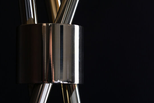 Abstract close-up photograph of a chrome metal connector joining tubular rods against a dark minimalist background, highlighting reflective surfaces, industrial design, texture and geometric form