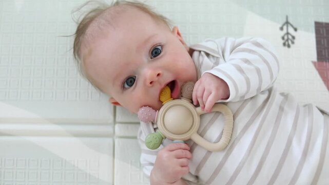 Close-up portrait of a baby chewing a silicone teething toy while lying on a soft blanket, soothing sore gums during teething in a bright nursery setting.