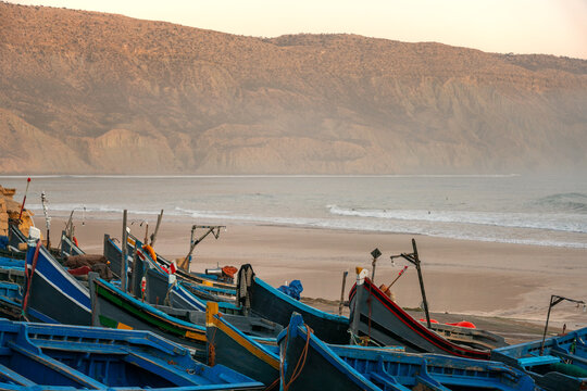 View of traditional blue fishing boats resting on the sandy beach at Imsouane with coastal cliffs and ocean waves in the background, Imsouane, Souss-Massa, Morocco.