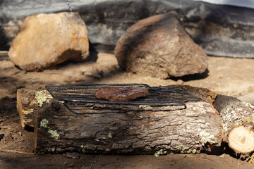 Fototapeta premium Cooking a rustic barbecue patty outdoors, grilling meat on an improvised campfire grill using logs and a metal grate in a natural setting