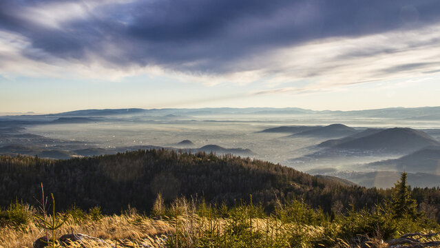 View of the Grazer Bergland mountains with a misty valley below, dense evergreen forests in the foreground, and a dramatic cloudy sky over Graz, Styria, Austria.