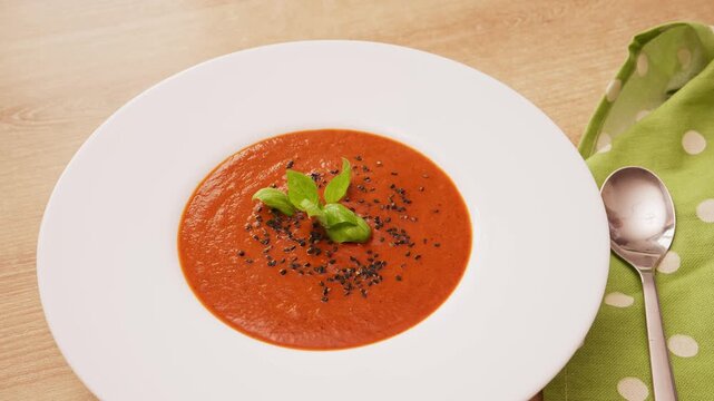 Hand placing fresh green basil leaf as garnish on top of hot red tomato and pepper cream soup in white ceramic bowl. Food styling for cooking vlog.