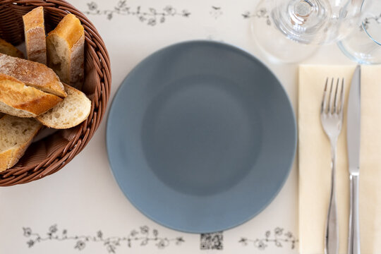 An empty blue-gray plate on a restaurant table setting, with a basket of sliced baguette bread, fork, knife, and wine glasses arranged around it, viewed from above.