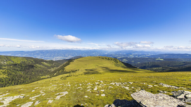 View of Koralpe mountain landscape with grassy slopes, rocky foreground, and distant mountain ranges under a clear blue sky in Weinebene, Styria, Austria.
