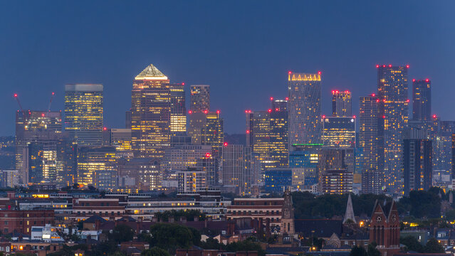 View of the illuminated Canary Wharf skyline at twilight with modern skyscrapers and office buildings rising above the surrounding residential area in London, England, United Kingdom.