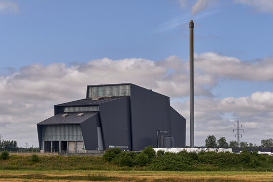 View of the modern biomass energy plant with its dark metallic facade, tall chimney, and stacks of wood fuel on the grounds in Esbjerg, Denmark.