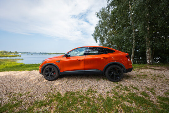 Renault Arkana RS Line coupe SUV in vivid orange parks by a calm lake. Birch trees line the right, black alloys and red calipers show. Wide angle side view in daytime.