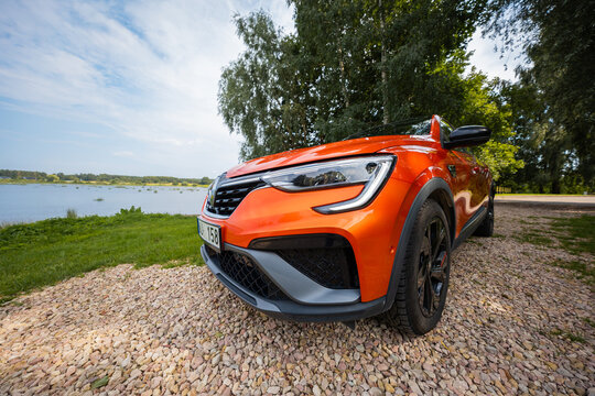 A vivid orange Renault Arkana RS Line stands on a gravel pull off by a calm lakeside, low wide angle view shows C shaped LED DRLs, black wheels, grille, and diffuser.