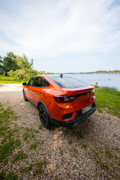 Orange Renault Arkana RS Line coupe SUV parks on gravel by a calm lake, rear three quarter view shows sloping roofline, black alloy wheels, RS Line badging, LED tail lamps.
