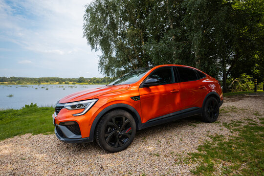 Renault Arkana RS Line in vivid orange sits on gravel by a calm lakeside, three quarters front left, showing RS badges, 18 inch alloys, red calipers, and black trim.
