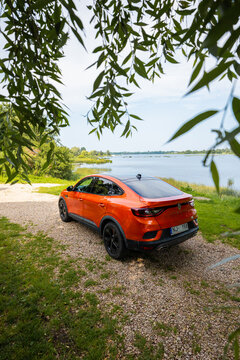 Renault Arkana RS Line in orange parks on gravel by a calm lakeside, willow leaves frame, daylight three quarter rear view shows black roof, diffuser, dual exhausts, red calipers.