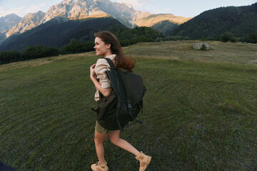 Obraz premium Cheerful woman outdoors, hiking with a backpack, grassy field and distant mountains in the background, sunlight on her hair, relaxed stride, natural colors and breeze