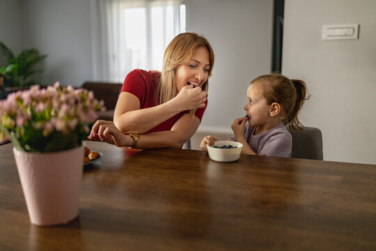 Mother and daughter sharing blueberries at kitchen table, family breakfast moment