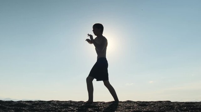 Young man on the seashore at sunset practices hand to hand combat and Muay Thai, throwing jabs, uppercuts and knee strikes. Shadow boxing. Solo training and preparation for youth competition.