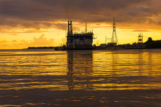 View of an industrial power plant facility on the water during a golden hour sunset with power lines and reflections in San Pedro de Macoris, San Pedro de Macoris, Dominican Republic.