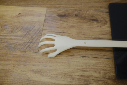 White plastic back scratcher with a claw shape lies flat on a light brown wooden surface in a quiet home interior during the daytime hours