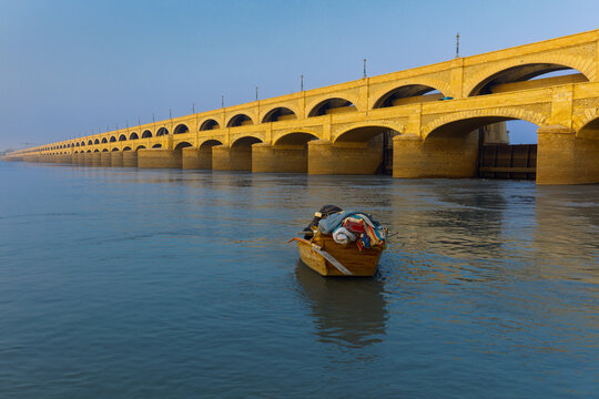 View of the historic Sukkur Barrage with its iconic stone arches spanning the Indus River, featuring a small wooden boat floating in the calm water in Sukkur, Sindh, Pakistan.