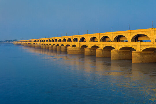 View of the historic Sukkur Barrage Bridge spanning the Indus River with its iconic yellow arches reflecting in the calm water under a clear blue sky in Sukkur, Sindh, Pakistan.
