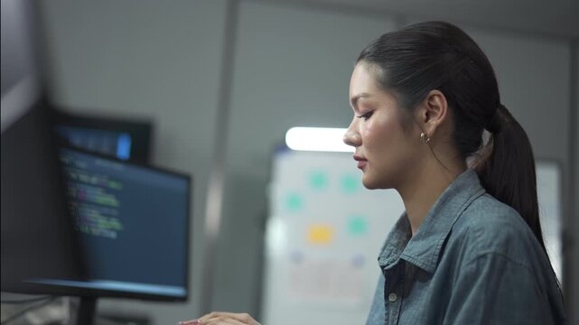 Young female software engineer writing program code on multiple desktop screens in a modern office.