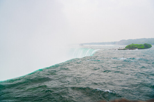 View of the powerful rushing water with mist rising over the edge and a small green island in the background in Niagara Falls, Ontario, Canada.