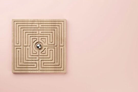 A miniature wooden maze puzzle with a metal ball in the center, presented on a soft pink background with ample copy space