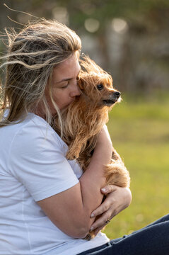 Young woman lovingly hugging a small dog in her lap; hair and fur blowing in the wind. Backlit by the sun in a park during summer.