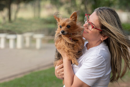 A young blonde woman wearing glasses, seen from the side, holding a small brown dog in a public park on a sunny day.