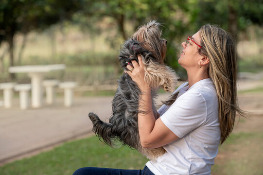 A young blonde woman wearing glasses, seen from the side, holding a Yorkshire Terrier facing her in a public park on a sunny day.