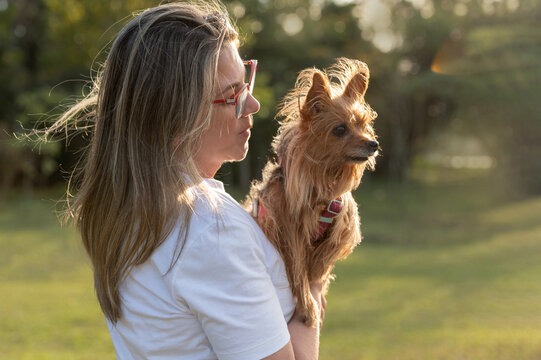 A young blonde woman wearing glasses, seen from the side, holding a small brown dog in a public park on a sunny day.