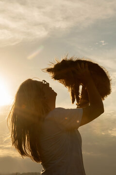 Silhouette of a woman lifting a small dog against the sunlight.