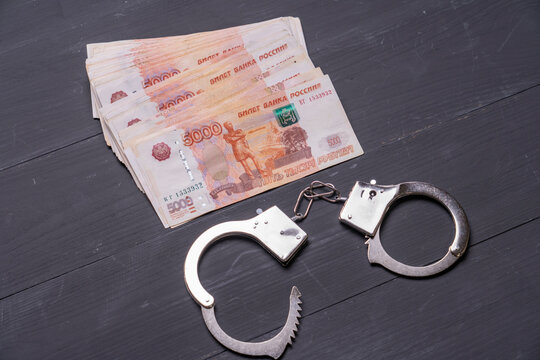 Russian 1000 ruble banknotes stack on dark wooden surface. Silver handcuffs lie beside the money. No people appear in the image. Suggests financial crime or police investigations