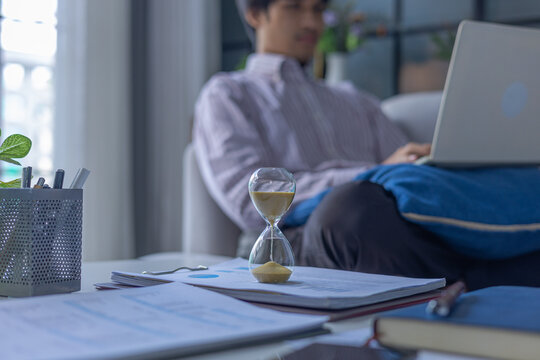 Man working on laptop sitting on sofa at home with running hourglass on the table. Concept for time management and extra over time working.