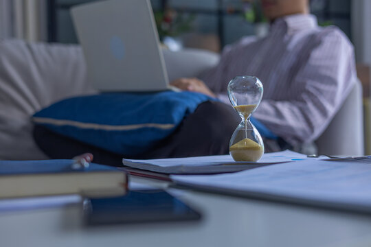 Selective focused on hourglass on the table with man working on laptop sitting on sofa at home. Concept for time management and extra over time working.