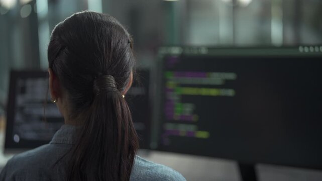 Rear view of young female software engineer writing program code on multiple desktop screens in a modern office.