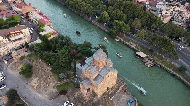 Panoramic aerial drone view of the historic orthodox Metekhi Church situated on a rocky cliff along the Kura River in Old Tbilisi, surrounded by cityscape and lush green trees, Georgia.