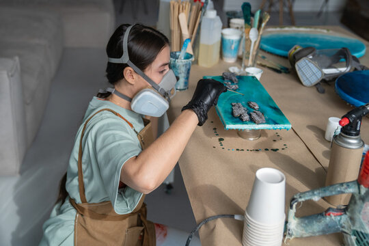 Talented woman artist creating liquid art painting, holding stick and adding drops of epoxy resin, focused female crafter in protective respirator and gloves dripping liquid resin over canvas surface.