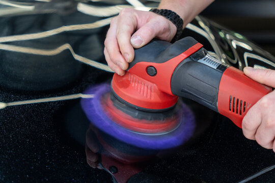 Close up of a professional car detailing process. Mans hands hold a red electric orbital dual action polisher machine with a purple microfiber wool pad buffer tool to polish a black auto paint hood. 
