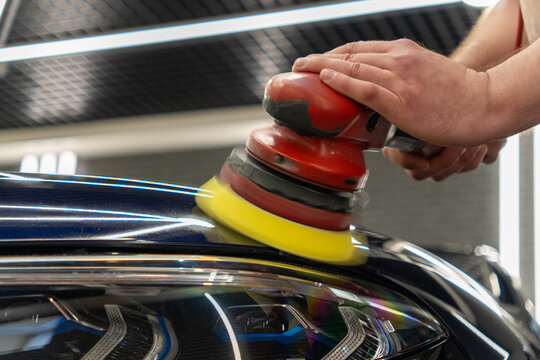 Professional car detailing process. Hands of a worker polishes the black paint of a luxury vehicle body using an electric orbital dual action polishing machine with a yellow foam pad buffer tool. 