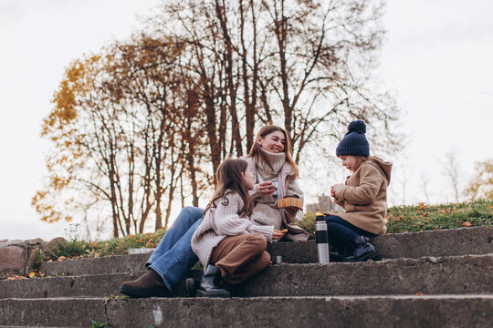 Happy young mother with daughters having picnic on concrete steps in autumn park. Family leisure, autumn outdoor activities, mother's day, togetherness, parenthood, seasonal lifestyle, warm clothing.