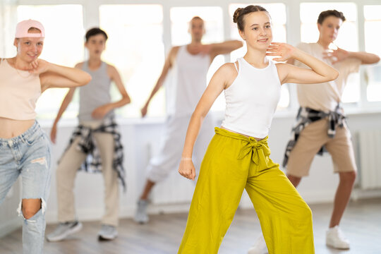Young girl in bright clothes, together with group of teenage friends, learn to dance hip hop in choreographic hall. Students move to modern music, do moonwalking, squats, bends, work with body